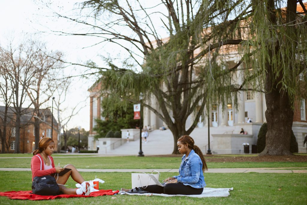 two students with electronic devices sitting on the grass in front of Gorgas Library